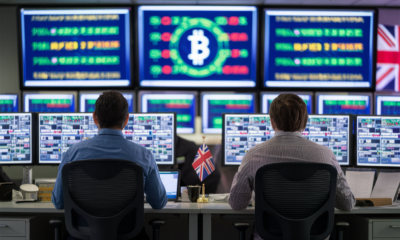 A large control room with several screens showing crypto financial symbols. Two office workers with their back to viewer sit at their desk. A UK flag is on the wall