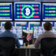 A large control room with several screens showing crypto financial symbols. Two office workers with their back to viewer sit at their desk. A UK flag is on the wall