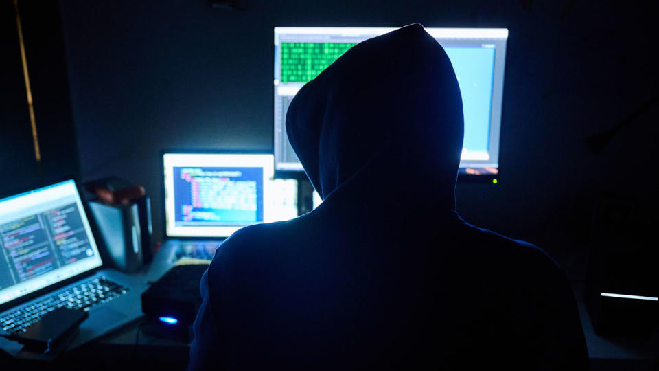 A man types at a computer as fake HTML pages and hacker programs appear on the screens in front of him.